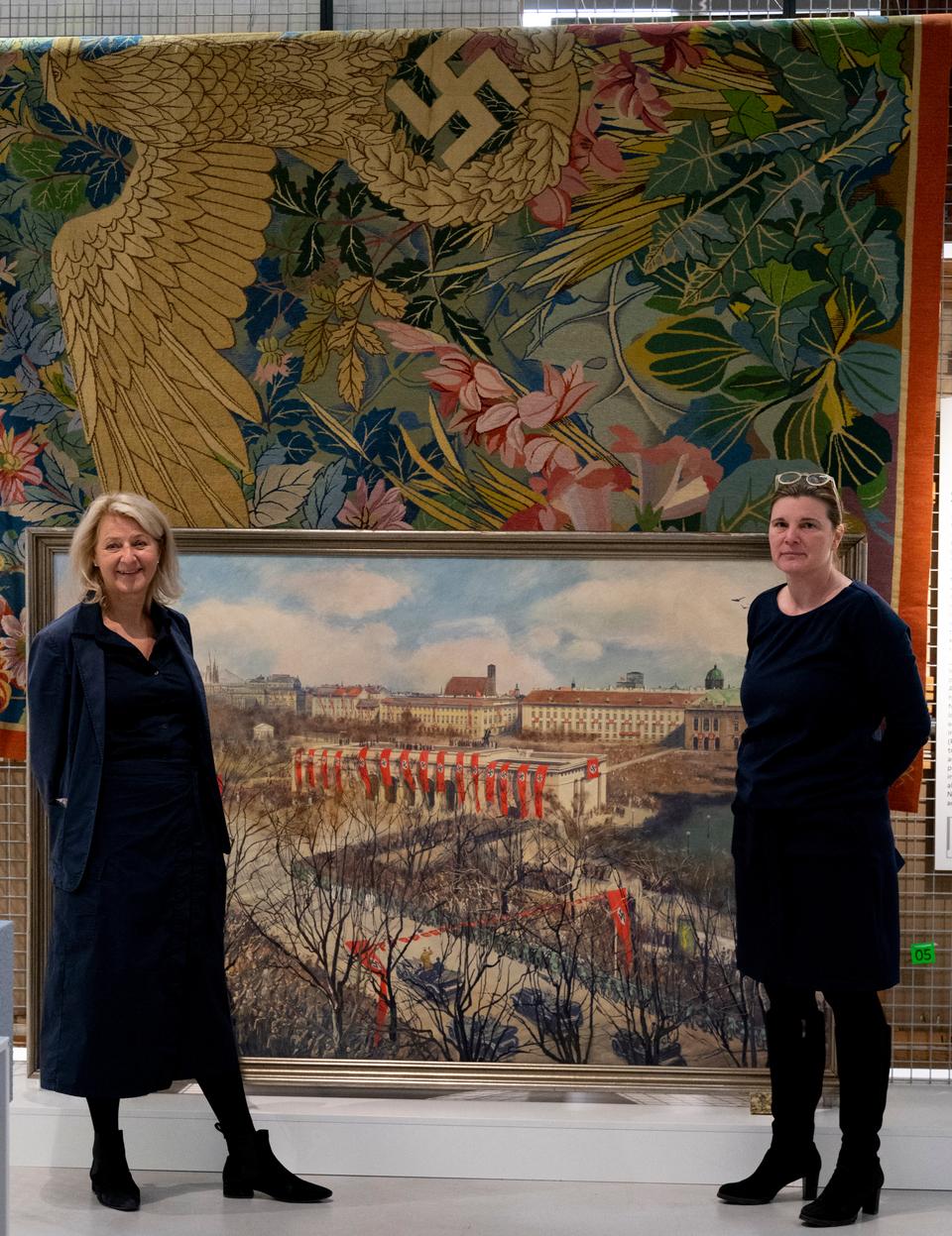 Curators Sabine Plakolm-Forsthuber (L) and Ingrid Holzschuh (R). Behind it is a painting of a Viennese opera adorned with a Nazi flag and a tapestry with a swastika.