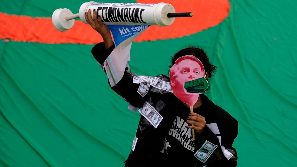 A demonstrator wears a mask depicting President Bolsonaro with a mouthful of US dollars, while holding a syringe representing Covid-19 vaccine, during a protest in Brazil on October 2, 2021.