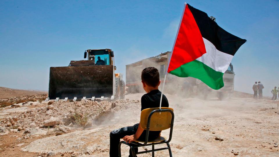 A Palestinian boy sits on a chair with a national flag as Israeli authorities demolish a school site in the village of Yatta, south of the West Bank city of Hebron and to be relocated in another area, on July 11 2018.