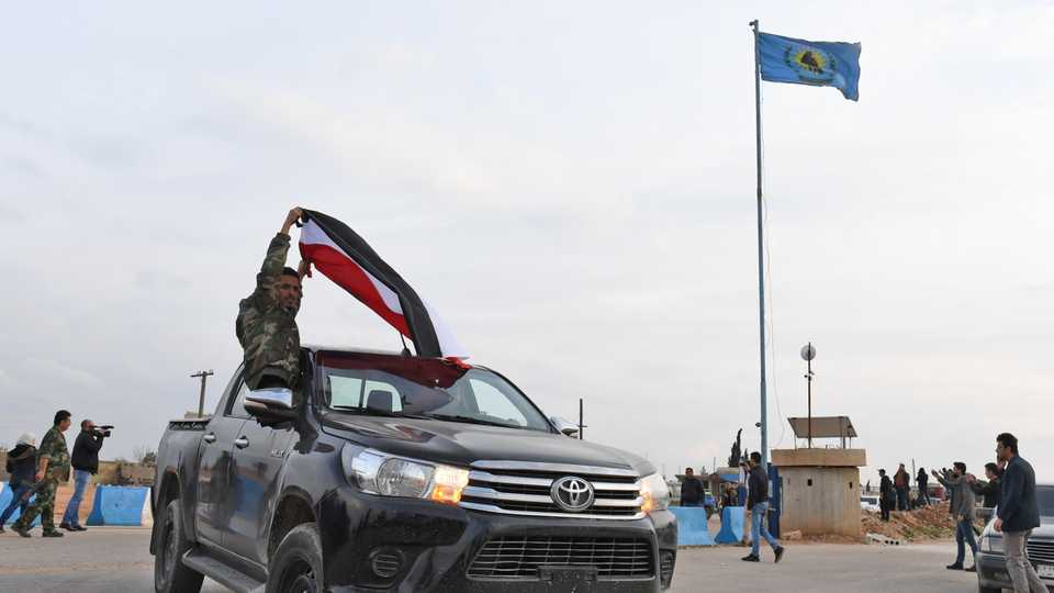Pro-Syrian regime fighters wave the regime flag as they ride on pickup trucks upon arriving in Syria's northern region of Afrin on February 20, 2018, with the YPG flag seen flying in the background.