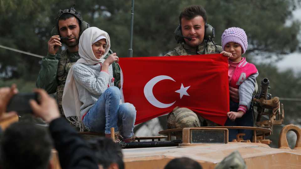 In this March 24, 2018 photo, Turkish soldiers atop a tank pose for pictures with Syrian children holding a Turkish flag in the northwestern city of Afrin, Syria.