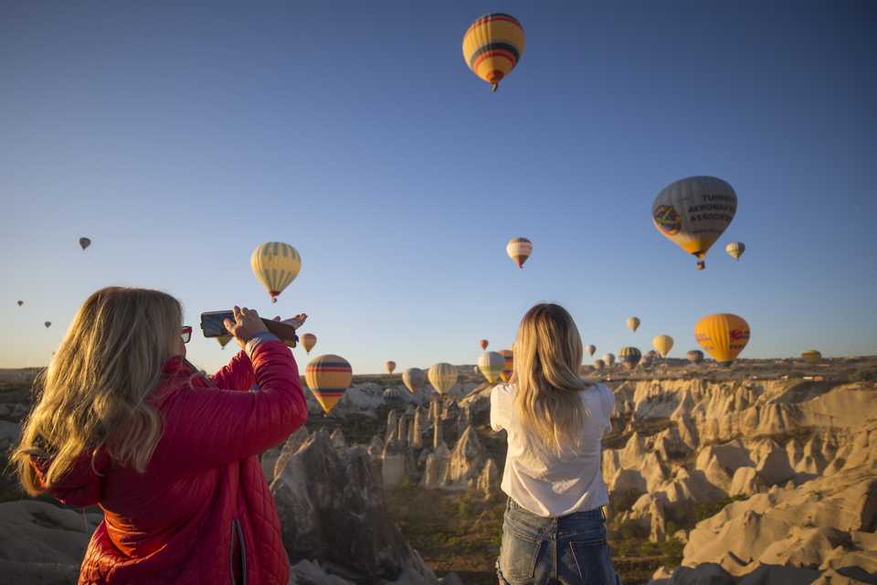 Tourists take photos as hot air balloons glide over Goreme district during early morning at the historical Cappadocia region, located in Central Anatolia's Nevsehir province, Turkey on September 30, 2019. Cappadocia is preserved as a UNESCO World Heritage site and is famous for its chimney rocks, hot air balloon trips, underground cities and boutique hotels carved into rocks.