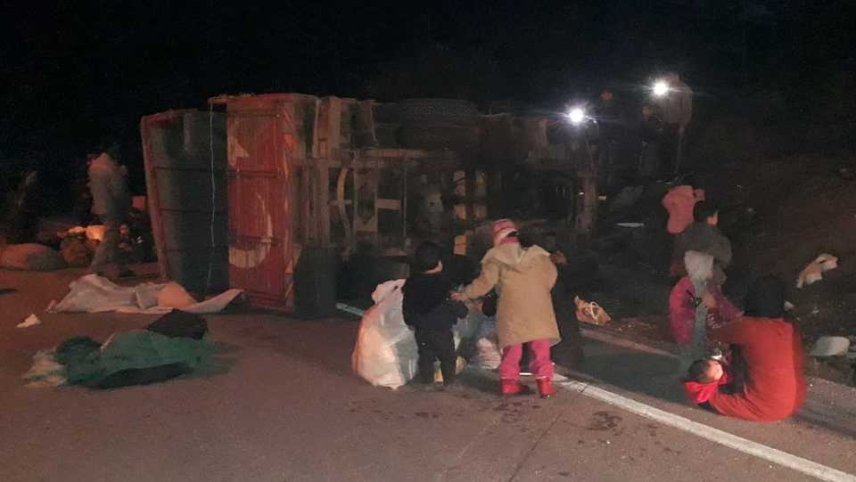 Irregular migrants stand at the roadside next to the overturned vehicle, December 26, 2019