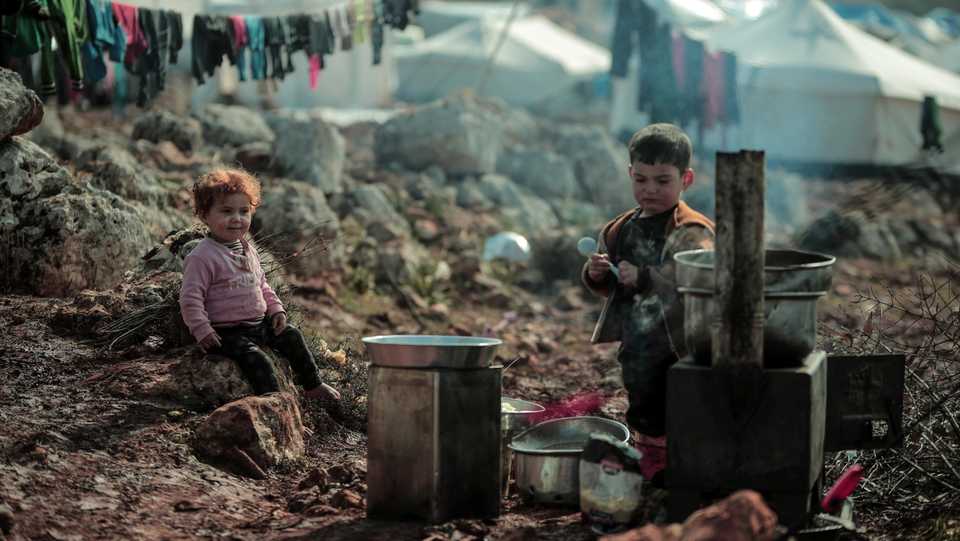 Two children wait for food in front of their tent in Harbnoush camp in harsh weather conditions.