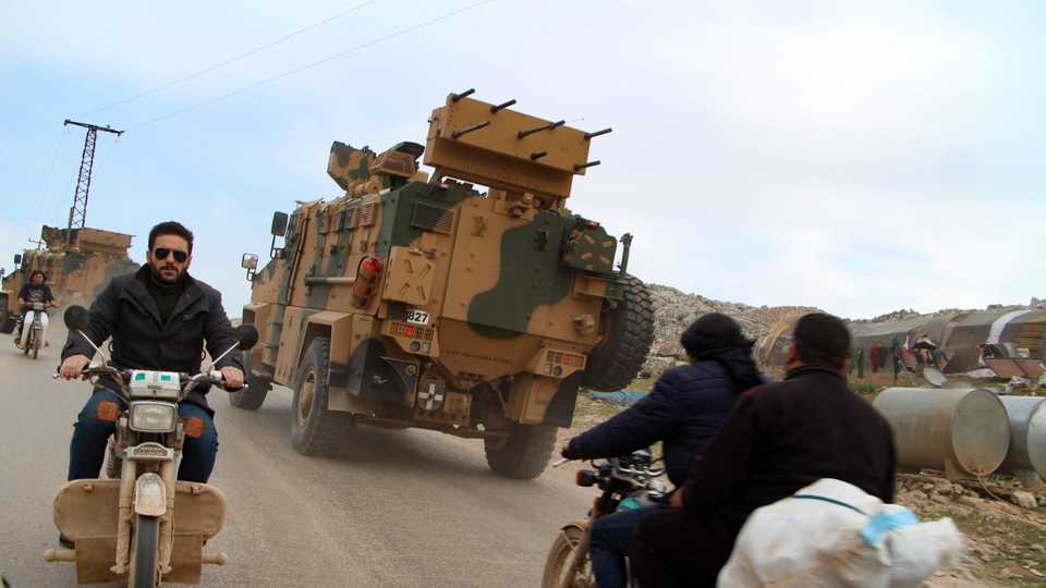 Syrians ride motorcycles as Turkish military vehicles drive past a camp for displaced people near the village of Kafr Lusin in Syria's northeastern Idlib province on March 6, 2020.