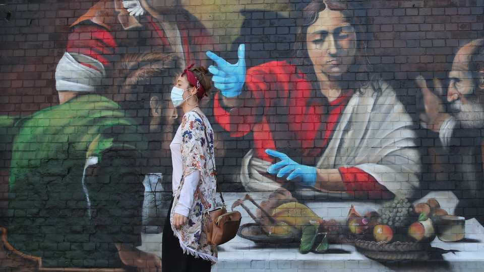 A woman stands in front of a mural by artist Lionel Stanhope depicting the painting ''The Supper at Emmaus' by Italian painter Caravaggio with the addition of blue nitrile gloves in south London, the UK on May 6, 2020.